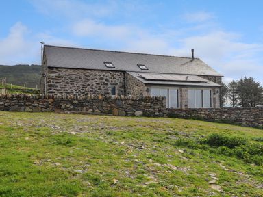 A stone house with a slate roof and solar panels behind a stone wall on a grassy slope at Beudy Mawr in Cwmystradllyn near Tremadog