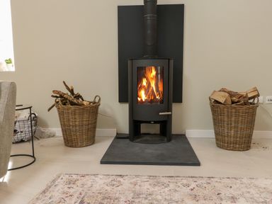 A wood stove with a fire burning two baskets of firewood on either side and a patterned rug in front at Beudy Mawr in Cwmystradllyn near Tremadog