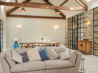 An open living and dining area with a beige sofa and pillows a wooden dining table with chairs and a vase with flowers a wooden cabinet stone wall and exposed wooden beams at Beudy Mawr in Cwmystradllyn near Tremadog