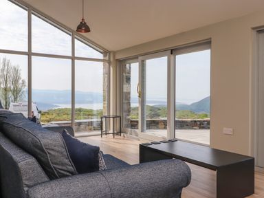 A living room with a blue sofa coffee table and round side table with large glass windows overlooking hills at Beudy Mawr in Cwmystradllyn near Tremadog