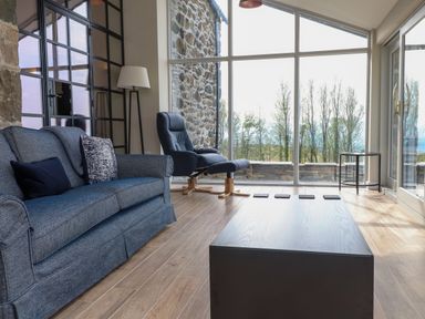 A living room with a blue sofa a black armchair a floor lamp and a wooden coffee table by large windows at Beudy Mawr in Cwmystradllyn near Tremadog
