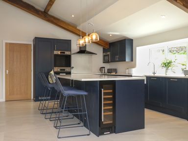 A kitchen with dark cabinets a central island with three bar stools pendant lights and a window with a vase of flowers at Beudy Mawr in Cwmystradllyn near Tremadog