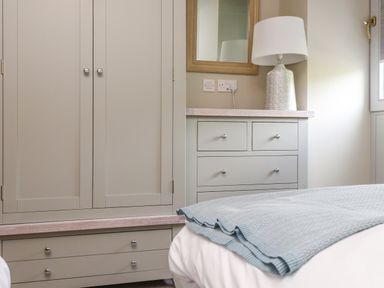 A bedroom corner with a grey wardrobe, chest of drawers, a lamp, a mirror, and part of a bed with a blue blanket at Beudy Mawr in Cwmystradllyn near Tremadog
