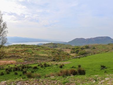 A grassy hillside with scattered bushes and a fence with mountains and a body of water in the background at Beudy Mawr in Cwmystradllyn near Tremadog