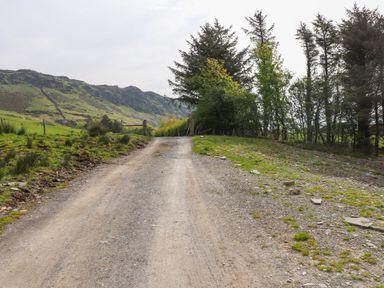 A gravel road leading towards a gate with trees on the right side and green fields on the left with hills in the background at Beudy Mawr in Cwmystradllyn near Tremadog