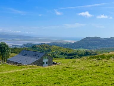 A stone house with a dark roof on a green hillside with hills and a body of water in the background at Beudy Mawr in Cwmystradllyn near Tremadog