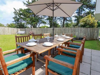 An outdoor dining area with a wooden table set with plates and glasses surrounded by wooden chairs with cushions under a large umbrella in a fenced garden