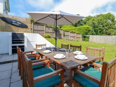 An outdoor dining area with a wooden table and chairs under a large umbrella next to a built-in barbecue and a fenced grassy yard at Surf Break Polzeath