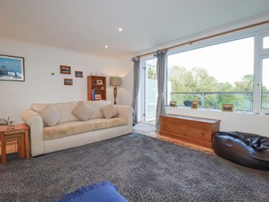 A living room with a beige sofa and striped cushions a wooden chest a bean bag and large window at Surf Break in Polzeath
