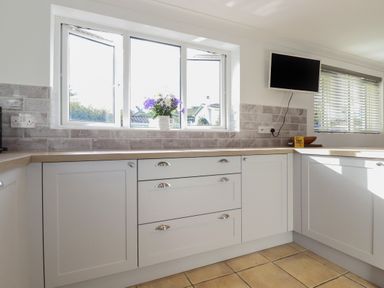 A kitchen with white cabinets a wooden countertop a flower vase on the windowsill a wall-mounted television and blinds on a window at Surf Break in Polzeath