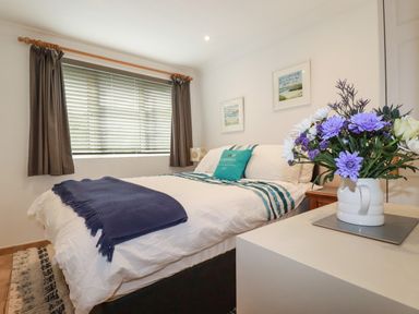 A bedroom with a bed covered in white and blue bedding a window with blinds and curtains and a vase of flowers on a white table at Surf Break in Polzeath