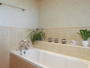 A bathroom with a white bathtub beige tiled walls plants and candles on a shelf at Surf Break in Polzeath