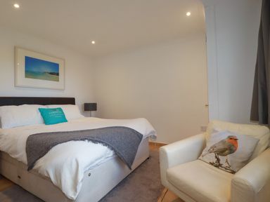 A bedroom with a bed a gray throw blanket a chair with a bird pillow and a framed picture on the wall at Surf Break in Polzeath