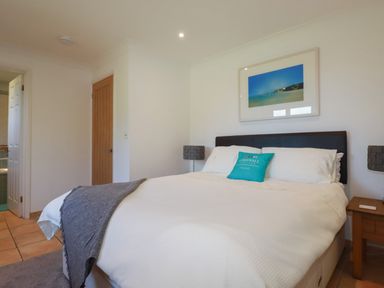 A bedroom with a bed with white bedding and a blue pillow a bedside table with a lamp a wooden door and a picture on the wall at Surf Break in Polzeath