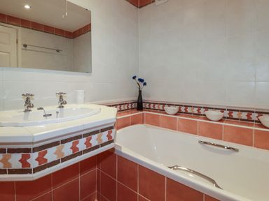 A bathroom with a bathtub and a sink with taps a mirror above and tiles with a patterned border at Surf Break in Polzeath