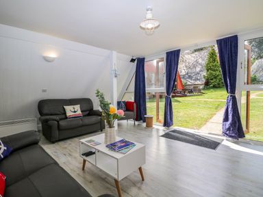 A living room with black sofas a white coffee table with magazines and flowers and large glass doors with blue curtains overlooking a garden at Chalet 58 - The Beach House in Kingsdown