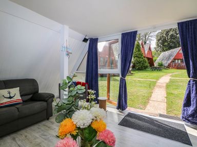 A living room with a black sofa and flowers on a table next to a glass door with blue curtains showing a garden outside at Chalet 58 - The Beach House in Kingsdown