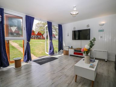 A living room with a coffee table with flowers a wall mounted TV and large glass doors with blue curtains at Chalet 58 - The Beach House in Kingsdown