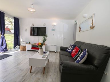 A living room with a black sofa with decorative pillows a white coffee table with flowers a wall-mounted TV and large windows with blue curtains at Chalet 58 - The Beach House in Kingsdown