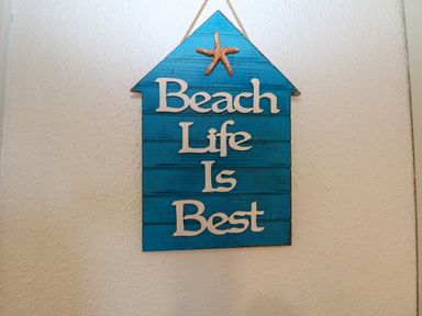 A blue wooden wall hanging shaped like a house with a starfish and the words Beach Life Is Best on a textured white wall at Chalet 58 - The Beach House in Kingsdown