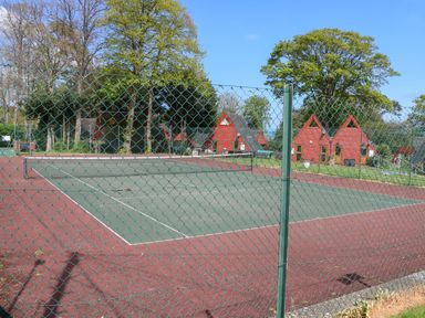 An outdoor tennis court with a green chain-link fence around it and red and green surface with red triangular houses and trees in the background at Chalet 58 - The Beach House in Kingsdown