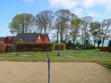 A sandy volleyball court with a net in front of red triangular cabins and trees by the sea at Chalet 58 - The Beach House in Kingsdown