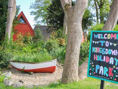 An outdoor scene with a small red and white boat used as a planter surrounded by greenery and a colorful welcome sign for Kingsdown Holiday Park