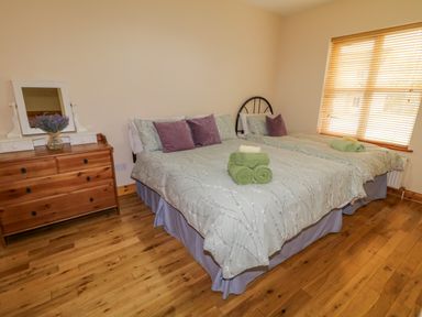 A bedroom with two beds covered in light bedding and purple pillows next to a wooden dresser with a mirror and flowers at 46 Dunalinn in Culdaff County Donegal