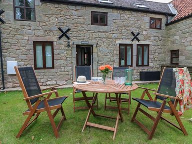 A wooden outdoor table with four chairs on grass in front of a stone building at The Haven in Norham