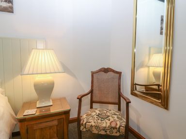 A wooden chair with a patterned cushion next to a wooden nightstand with a lamp and a coaster and a wall mirror at The Haven in Norham