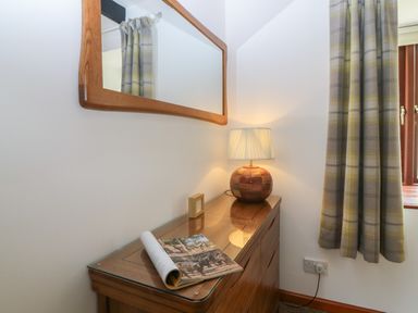 A wooden dresser with a table lamp a magazine and a small framed picture next to a window with plaid curtains at The Haven in Norham
