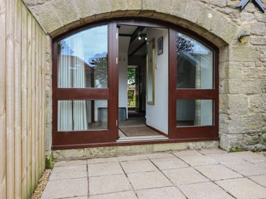 Arched wooden and glass double doors open to a hallway with a mirror on the wall and view of outdoor chairs at The Haven in Norham