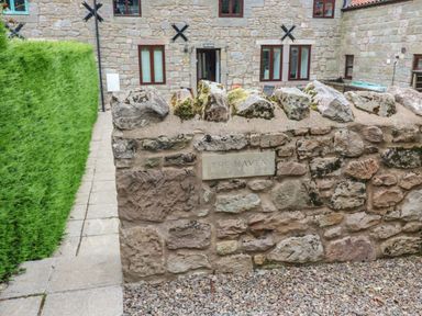 A stone wall with a plaque reading The Haven and a path with green shrubs leading to a stone building at The Haven in Norham
