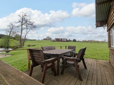 A wooden deck with a table and chairs overlooking a grassy field with cabins and trees at Hollie in Raglan