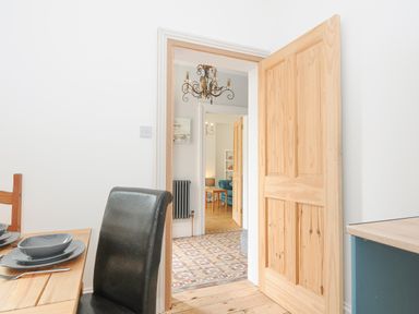 A dining area with a wooden table set with gray dishes and a black chair and an open wooden door showing a hallway with patterned flooring and a chandelier and a living room with a blue sofa at Albert House in Looe