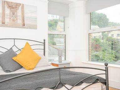 A bedroom with a metal bed frame, two colored pillows, a gray blanket, a side table with a lamp, and large windows overlooking houses and trees at Albert House in Looe