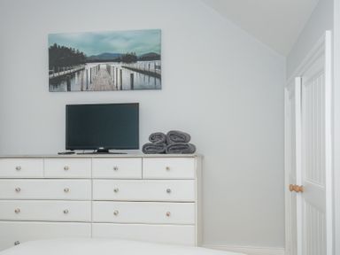 A bedroom with a white dresser holding a television and folded towels with a landscape painting on the wall at Albert House in Looe