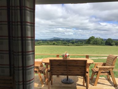 A wooden outdoor table with chairs and a flower basket on a deck overlooking a green field and distant hills at Poppy in Raglan