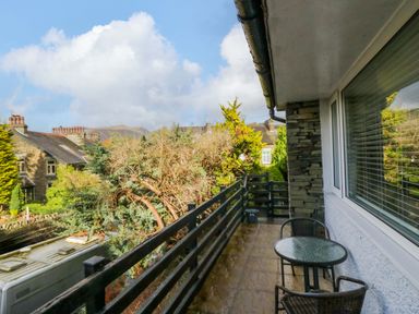 A small balcony with a glass round table and two chairs overlooking trees and neighboring houses at Lyndhurst in Ambleside