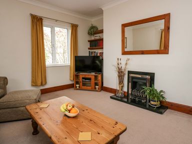 A living room with a wooden coffee table holding a bowl of fruit a sofa window with curtains a TV on a wooden stand fireplace with plants and a wooden framed mirror at Lyndhurst in Ambleside