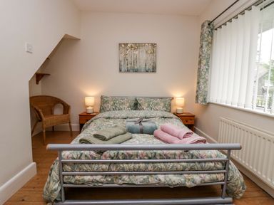 A bedroom with a double bed with floral bedding two bedside tables with lamps a wicker chair and a window with vertical blinds and floral curtains at Lyndhurst in Ambleside