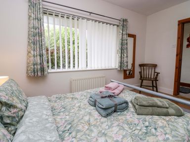 A bedroom with a bed with floral bedding folded towels on the bed a window with vertical blinds floral curtains a wooden chair and a wall mirror at Lyndhurst Ambleside