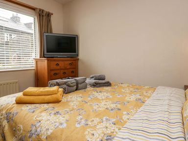 A bedroom with a bed covered in patterned bedding towels on the bed a wooden chest of drawers with a television on top and a window with blinds and curtains at Lyndhurst in Ambleside