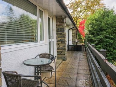 A narrow outdoor patio with a small round glass table and two wicker chairs along a white building with stone columns and a wooden fence surrounded by trees at Lyndhurst in Ambleside