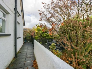 An outdoor pathway along a white building with a black rain gutter and railing overlooking a garden area with trees and patio furniture at Lyndhurst in Ambleside
