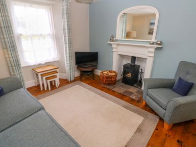 A living room with a grey sofa and armchair a fireplace with a stove and a TV on a small table at Orielton Cottage in Dinas Cross