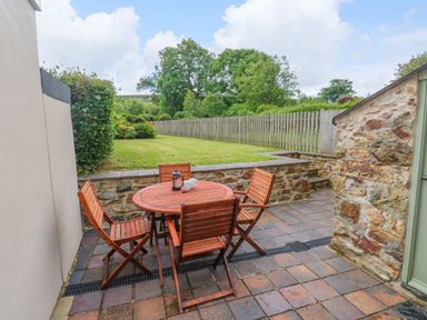 An outdoor patio with a round wooden table and four wooden chairs next to a stone wall and a garden with grass and trees at Orielton Cottage in Dinas Cross