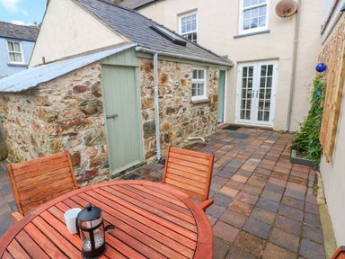 An outdoor patio with a wooden table and chairs next to a stone building with a green door and a building with white double doors at Orielton Cottage in Dinas Cross