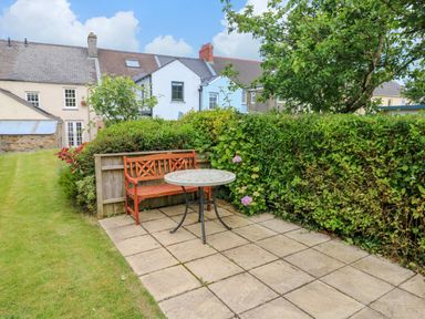 A garden patio with a wooden bench and round table near hedges and houses in the background at Orielton Cottage in Dinas Cross