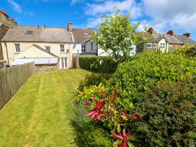 A backyard garden with green grass, bushes, and trees behind houses at Orielton Cottage in Dinas Cross